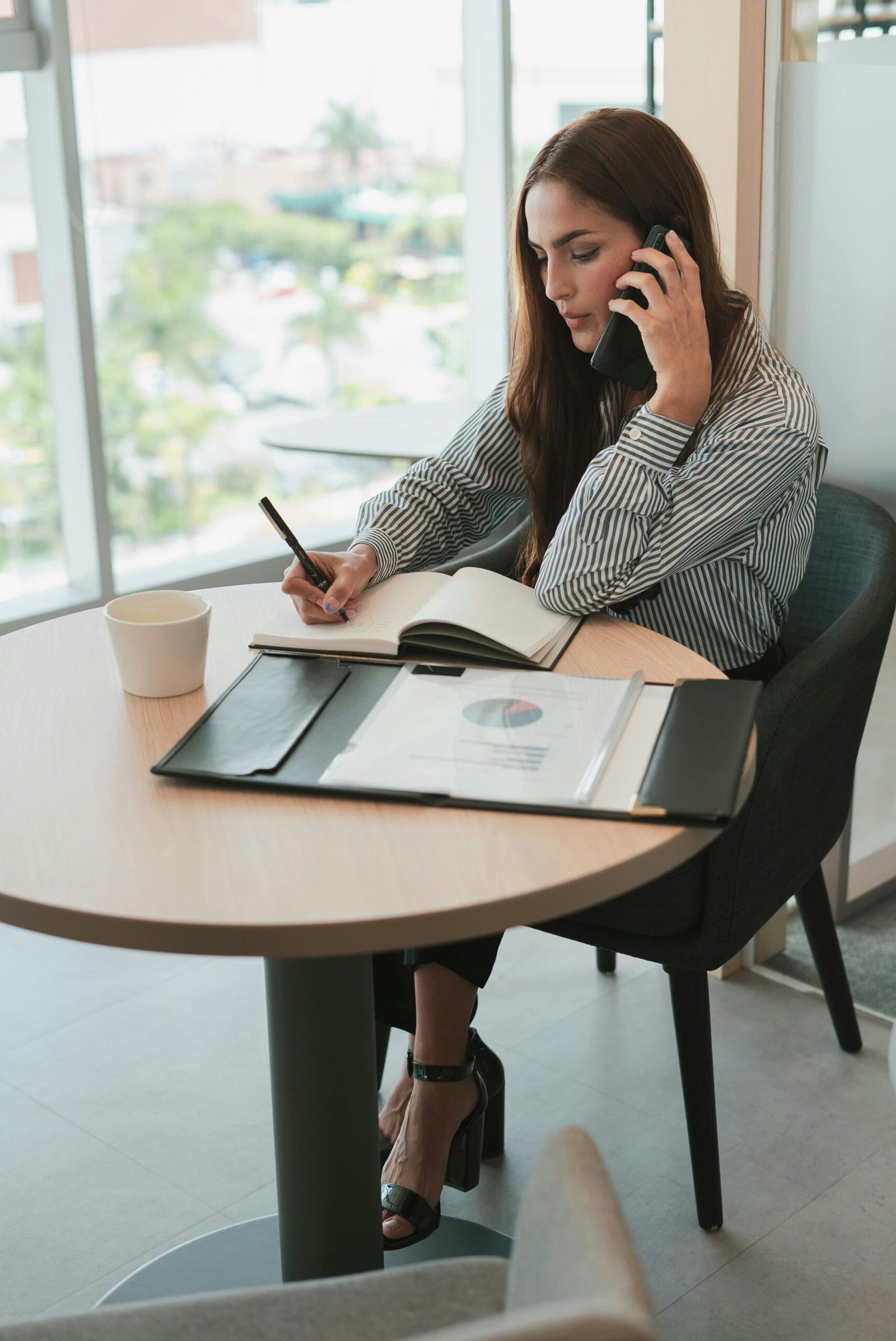 pexels-photo-10041241-10041241 Woman in striped shirt multitasking in an office, writing notes while on a phone call, business setting.